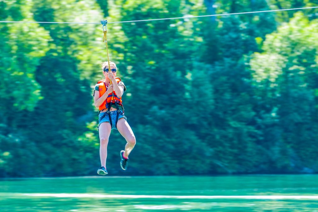 young girl zip lining over clear water, with large trees in the background