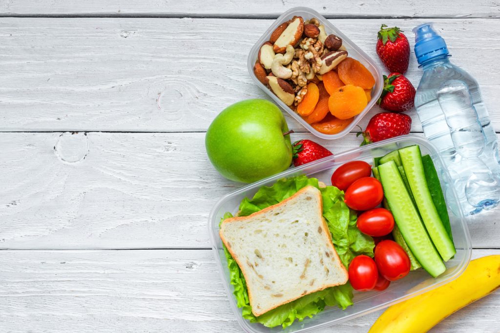 school lunch boxes with sandwich and fresh vegetables, bottle of water, nuts and fruits on white wooden background