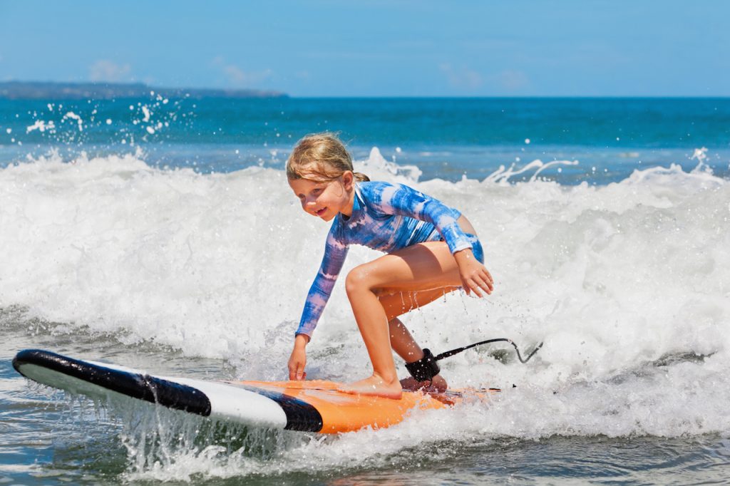 young surfer ride on surfboard with fun on sea waves. Active family lifestyle, kids outdoor water sport lessons and swimming activity in surf camp. Summer vacation with child.