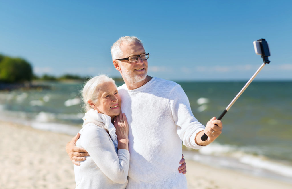 happy senior couple with smartphone selfie stick photographing and hugging on summer beach